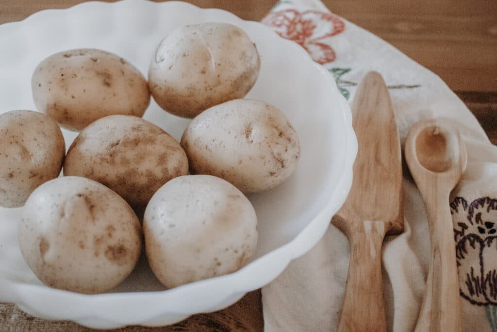 Whole potatoes in a white bowl next to a vintage embroidered tea towel and wooden utensils, preparing to make Best Roasted Garlic Mashed Potatoes.
