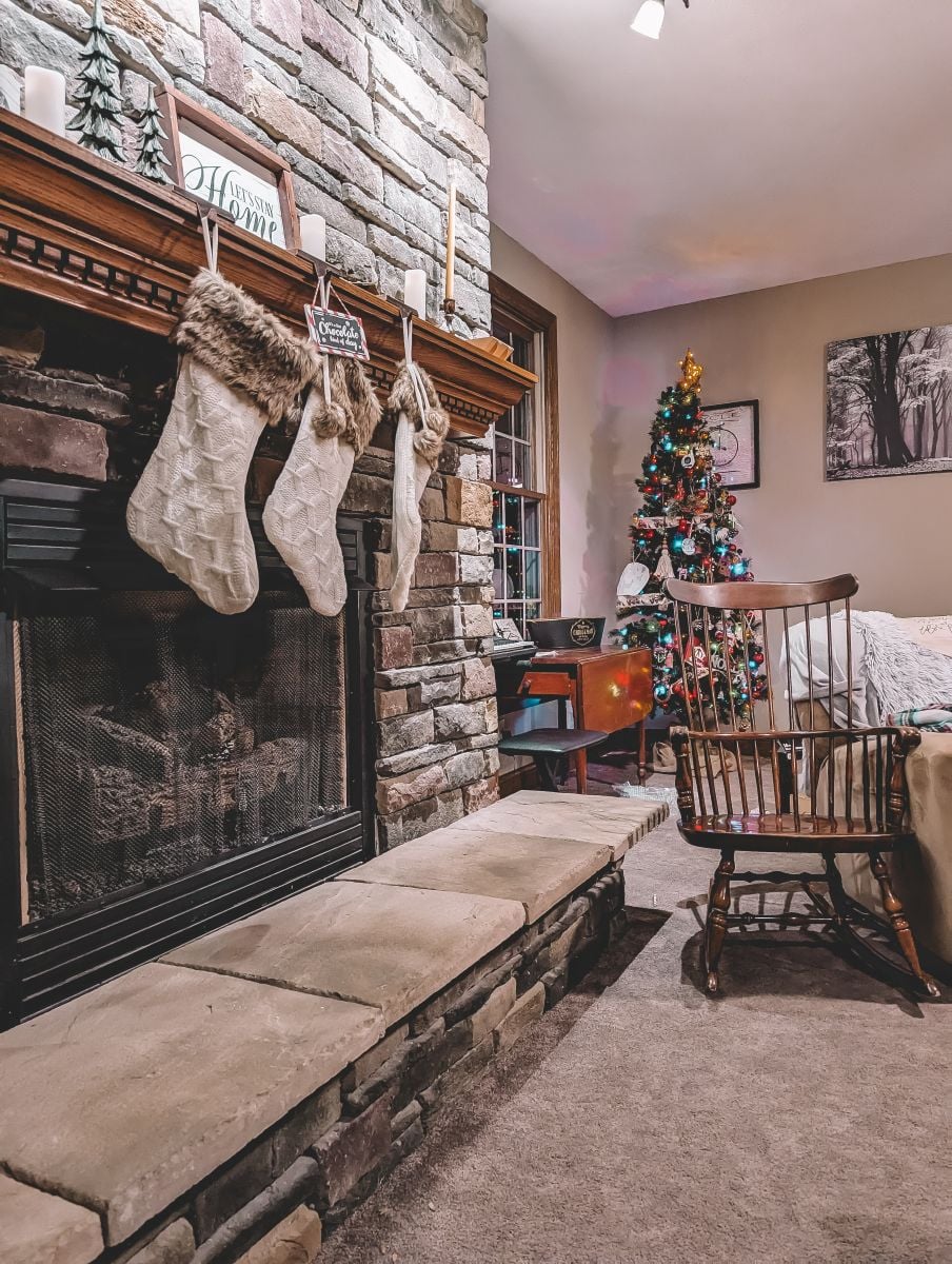 living room with a stone fireplace. three christmas stockings are hanging from a wooden mantle. in the far corner is a lighted and decorated christmas tree a wooden rocking chair and a vintage wooden table with a black and gold mental basket that says Merry Christmas