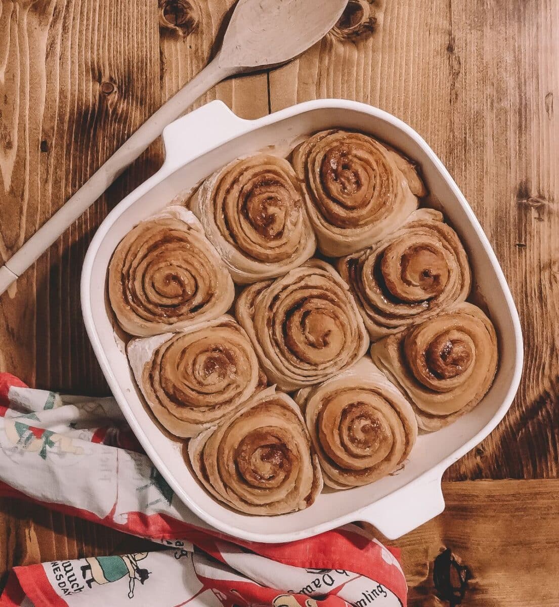 a white dish with cinnamon rolls inside sitting on a wooden table beside a wooden spoon and a red white and green kitchen towel