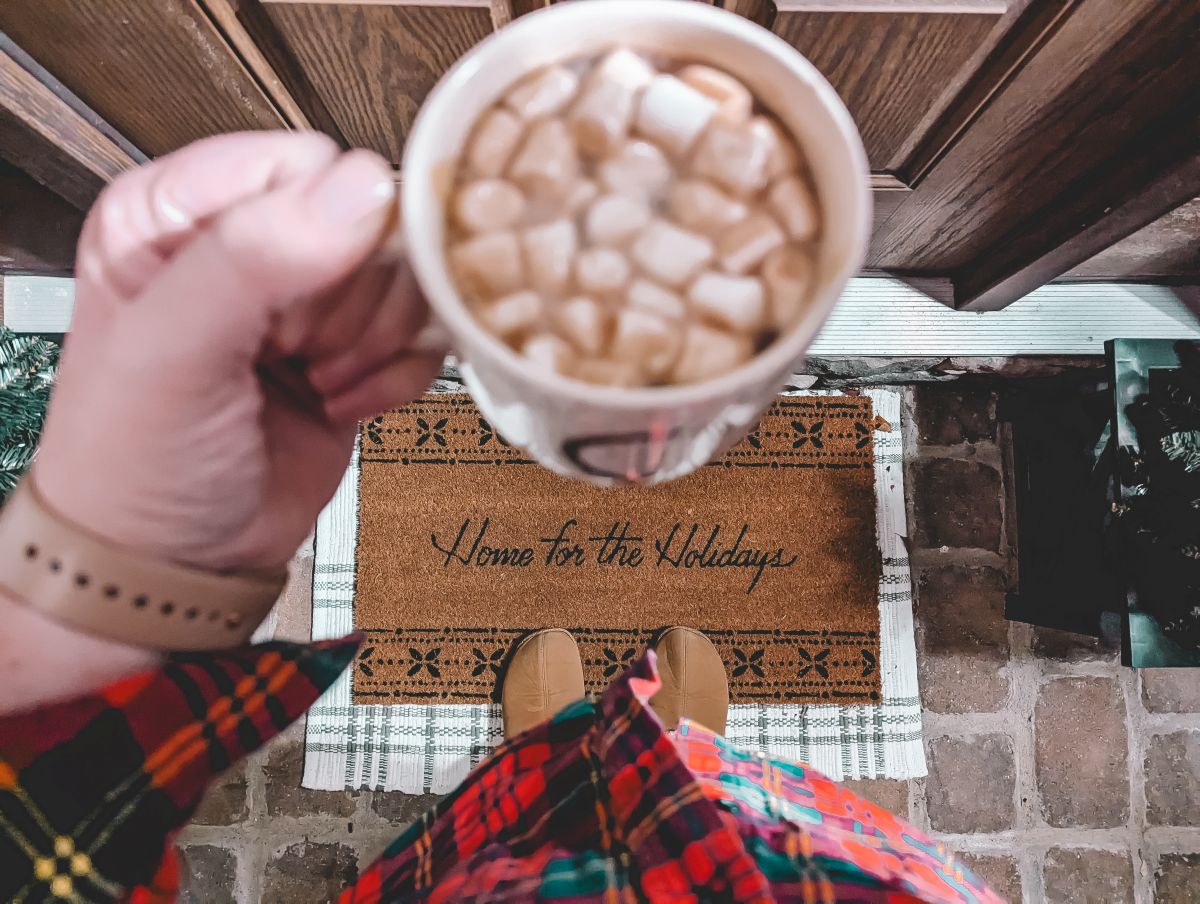 left hand of a caucasian woman holdind a coffee mug with hot cocoa and mini marshmallows in it. below is a porch rug that reads Home for the Holidays. In view is the woman's red and green flannel shirt and the brick paving stones of the porch